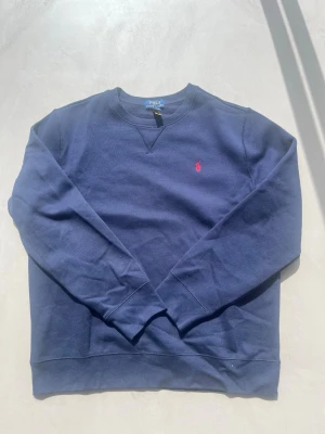 Marinblå sweatshirt från Polo Ralph Lauren - Klassisk marinblå sweatshirt från Polo Ralph Lauren med rund halsringning och röd broderad logga på bröstet. Tröjan har långa ärmar och ribbade muddar vid ärmslut och nederkant. Perfekt för en avslappnad och stilren look. Rekommenderas för dem som är 163-174 cm lång.