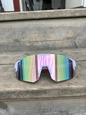 Pastellrosa vit ram Sportiga regnbågsfärgade solglasögon - Säljer ett par sportiga solglasögon med spegelglas i regnbågsfärger och bred, kantig form. Perfekta för dig som vill sticka ut med en futuristisk look. Lätta bågar i syntetmaterial och stor lins som täcker bra.