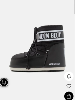Svarta Moon Boot Classic boots - Säljer ett par svarta Moon Boot Classic boots med vit logga och snörning framtill. Skorna har en vadderad och hög design med vit sula och är perfekta för kalla dagar. Materialet är syntet och textil, vilket gör dem både varma och bekväma. 