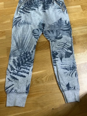 Grå mjukisbyxor med bladprint 157 - Snygga grå mjukisbyxor från 157 med mörkblått blad- och ormbunksmönster. Byxorna har muddar vid bensluten och dragkedjor på fickorna. Perfekta för chill dagar och riktigt skön passform.