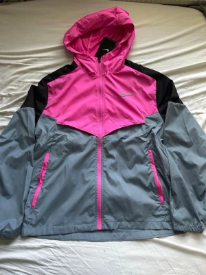 Nike Miler Rosa - Nike Storm-FIT Miler vindjacka i färgerna rosa, grå och svart. Jackan har huva, hel dragkedja och två fickor med rosa detaljer. Tillverkad i lätt och vattenavvisande material, perfekt för blåsiga dagar. Snygg sportig look med Nike-logga på bröstet.