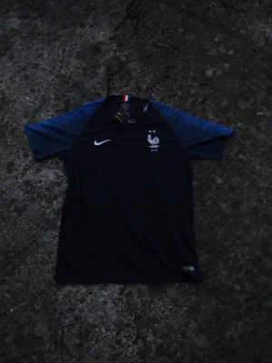 Frankrike fotbollströja från 2018 - Mörkblå fotbollströja från Nike med Frankrikes landslagströja från 2018. Ärmarna är i en ljusare blå nyans och har en fransk flaggdetalj i nacken. Tillverkad i lätt och ventilerande material, perfekt för träning eller match.