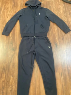Mörkblå Polo Ralph Lauren set - Mörkblått set från Polo Ralph Lauren med dragkedja, huva och två fickor framtill. Klassisk vit logga broderad på bröstet. Perfekt för en avslappnad och sportig stil. Tillverkad i mjukt bomullsmaterial.