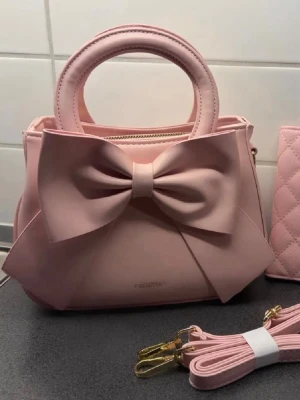 Rosa Väska - Supersöt rosa väska med en stor rosett på framsidan och rundade handtag. Väskan stängs med dragkedja och har 3 fack (se bild 2). Jätte girly och cute 🥰