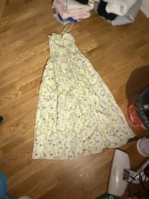 Ljusblommig långklänning med smala axelband - Söt långklänning med smala axelband och blommigt mönster i ljusa toner. Klänningen har en smockad överdel och faller fint ner mot golvet. Perfekt för varma dagar och somriga tillfällen.