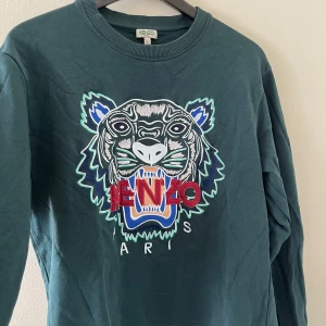 Kenzo sweatshirt med tigerbrodyr - Mörkgrön sweatshirt från Kenzo med ikonisk tigerbrodyr och röd/blå/vit Kenzo-logga framtill. Tröjan har rund hals, långa ärmar och är gjord i mjuk bomull. Perfekt statement-plagg för dig som gillar streetstyle och vill sticka ut. Snygg att dressa med en skjorta under! Nypris: 3999kr