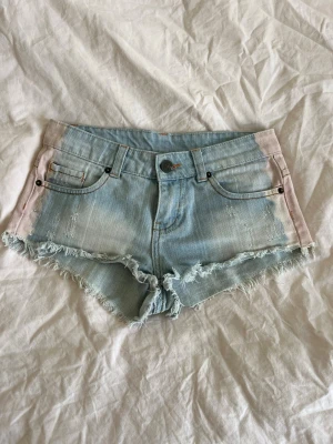 Ljusblå fransiga jeansshorts - Säljer ett par ljusblå jeansshorts med slitna detaljer och fransig kant. Shortsen har fem fickor, knappgylf och en cool tvätt med svagt rosa toner på sidorna. Perfekta för varma dagar och en avslappnad stil.