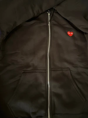 Svart hoodie Comme des Garçons Play - Svart hoodie med dragkedja från Comme des Garçons Play. Klassisk design med röd hjärtlogga med ögon på bröstet, huva och känguruficka framtill. Tillverkad i mjukt bomullsmaterial som känns skönt mot huden. Perfekt för en clean och stilren look.