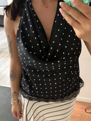 Svart prickig halterneck topp med spets - Snygg svart halterneck topp med vita prickar och öppen rygg. Toppen har en lös passform och spetsdetalj längst ner. Perfekt för dig som gillar en unik och trendig look.