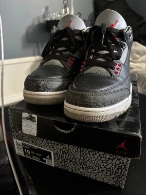 Air Jordan 3 Retro Black Cement - Säljer ett par Air Jordan 3 Retro Black Cement sneakers med svart och grått läder, ikoniskt elefantmönster på tå och häl, röda detaljer och vita sulor. Klassisk Jumpman-logga på plösen och svarta skosnören. Perfekt för dig som gillar streetwear och sneakerskultur.