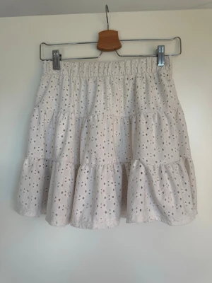 Vit volangkjol med broderade detaljer - Söt kortkjol i vitt med broderade hålmönster och volanger. Kjolen har resår i midjan för skön passform och är perfekt för sommarens alla outfits. Tillverkad i lätt bomullstyg som känns luftigt och fräscht.