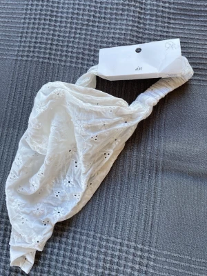 Vit broderad hårband från H&M - Säljer ett vitt hårband från H&M med broderade blommor och hålmönster. Hårbandet har bred form och resår baktill för skön passform. Perfekt accessoar för att lyfta din look med en romantisk touch.