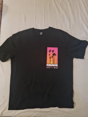 Svart Miami t-shirt med tryck - Cool svart t-shirt med färgstarkt Miami-tryck framtill i rosa, orange och gult. Motiv med palmer och texten 'Miami Florida' ger en skön sommarkänsla. Klassisk passform och rund hals. Perfekt för dig som gillar streetwear och snygga prints.