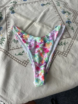 Färgglad blommig bikiniunderdel - Säljer en bikiniunderdel med blommigt mönster i rosa, lila, grön, gul och blå toner. Modellen har hög skärning och smal passform, perfekt för sommaren. Materialet känns mjukt och stretchigt, perfekt till sommaren. Oanvänd