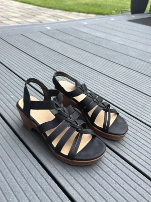 Svarta kilklackssandaler från Alley comfort i strl 39 - Snygga svarta sandaler från Alley med flätade remmar och öppen tå. Sandalerna har en bekväm kilklack i trälook och elastiska band för skön passform. Perfekta för dig som vill ha både stil och komfort under sommaren.