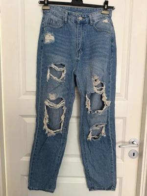 Ljusblå ripped mom jeans - Säljer ett par ljusblå mom jeans med stora slitningar och hål på båda benen. Jeansen har hög midja, raka ben och klassisk femficksmodell. Tillverkade i denim med en cool vintage vibe och riktigt trendig look.