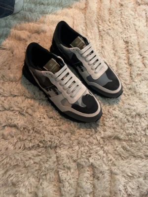 Valentino camo sneakers svart/vit - Snygga sneakers från Valentino med svart, grå och vit camo-mönstrad ovandel. Skorna har vita snören, svart sula och detaljer i mocka och textil. Perfekta för dig som vill sticka ut med en unik och trendig look. Storlek 43, de var för små för mig så väljer att sälja vidare.