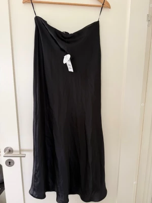 Svart satinkjol från Next  - Elegant svart satinkjol från Next i storlek 38/M. Kjolen är helt oanvänd och har fortfarande prislappen kvar. Jag köpte den men fick aldrig något tillfälle att använda den, så den är i nyskick.  Kjolen sitter fint och tajt i midjan, medan den faller ut något längre ner för en mjuk och feminin silhuett. Det glansiga satinmaterialet ger ett stilrent och elegant intryck som passar både till vardag och fest.