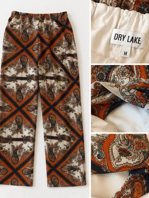 Mönstrade mjukisbyxor från Dry Lake - Säljer ett par mjukisbyxor från Dry Lake med paisleymönster i orange, svart och beige. Byxorna har rak passform, resår i midjan och är tillverkade i ett mjukt, glansigt material som känns lyxigt. Perfekta för dig som vill sticka ut med coola detaljer.