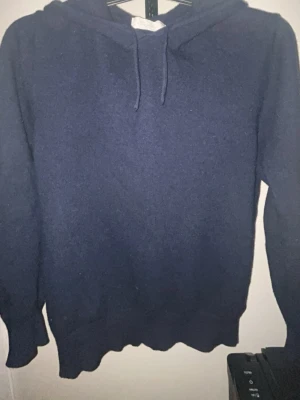 Kashmir hoodie - En enkel och stilren mörkblå kashmir hoodie med huva och snören. Hoodien har långa ärmar och ribbade muddar vid ärmslut och nederkant. Perfekt för en avslappnad och bekväm stil.