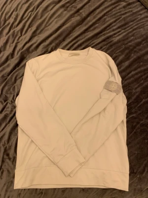 Stone Island ” ghost piece ”  - Säljer en stilren vit sweatshirt från Stone Island med den klassiska logotypen på vänster ärm. Tröjan har rund halsringning, långa ärmar och ribbade muddar. Perfekt för dig som gillar minimalistisk och clean streetwear.