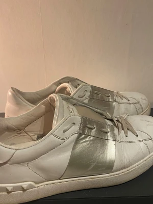 Vita och silverfärgade sneakers från valentino  - Snygga sneakers från Valentino i vitt läder med en bred metallic-silver panel på sidan. Klassisk låg modell med vita snören och diskret branding på plösen. Perfekta för dig som vill ha en stilren men ändå unik look.
