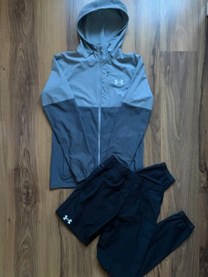 Under Armour träningsset grå/svart - Snyggt träningsset från Under Armour med en tvåfärgad grå och svart jacka med huva och dragkedja samt matchande svarta byxor. Båda är storlek S. 