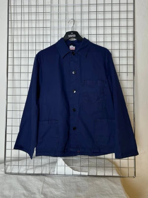 Worker jacka - Stilren marinblå overshirt från Le Mont Saint Michel med tre utanpåliggande fickor och klassisk krage. Jackan har knäppning framtill med svarta knappar och raka, långa ärmar. Perfekt för lager på lager och enkel att matcha.