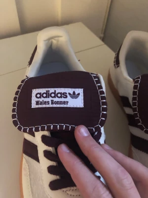 Adidas Wales Bonner sneakers vita/lila - Säljer ett par Adidas x Wales Bonner sneakers i vitt och mörklila med klassiska tre ränder på sidan och kontrastsömmar runt plösen. Skorna har en gummisula och är tillverkade i läder och mocka. Perfekta för dig som gillar unika samarbeten och snygg retrostil.