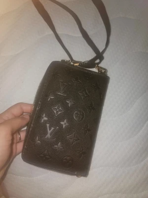 svart plånbok med axelrem - Snygg svart plånbok i präglat läder med klassiskt monogrammönster. Plånboken har dragkedja runtom och en avtagbar axelrem, samt flera fack för kort och mynt. Perfekt för dig som vill ha både stil och funktion.