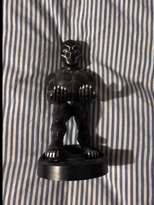 Black Panther figurhållare Marvel - Cool Black Panther figurhållare från Marvel i svart plast med lila detaljer. Figuren står på en rund bas och har utsträckta armar för att hålla till exempel en spelkontroll eller mobil. Perfekt för Marvel-fans som vill ha snygg förvaring.