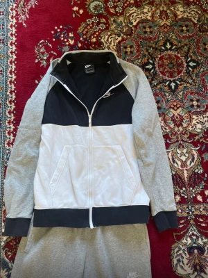 Nike grå och vit dress set - Nike loungewear set med ziptröja och byxor i grått, vitt och svart. Tröjan har dragkedja, fickor och svart panel upptill. Byxorna är grå med vita detaljer på benen och har resår i midja och benslut. Perfekt för chill dagar hemma.