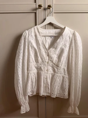 Vit broderad blus från Bubbleroom - Superfin vit blus från Bubbleroom med broderade hål och v-ringning. Blusen har markerad midja, volang nertill och långa ärmar med resår vid ärmslut. 