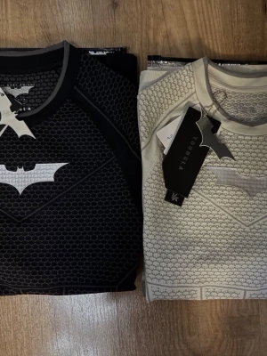 Batman T-shirt från YoungLA bytes mot skor eller kläder - Två snygga Batman-inspirerade t-shirts från YoungLA, en i svart och en i beige. Båda har ett coolt bikakemönster och en stor Batman-logga på bröstet. T-shirtsen har rund hals och korta ärmar, perfekta för dig som vill sticka ut. Finns inge taggar kvar