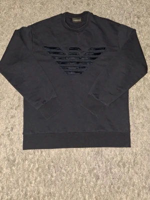 Svart sweatshirt från Emporio Armani - Mörkblå sweatshirt från Emporio Armani med det ikoniska örnlogotypen i sammet på bröstet. Tröjan har rund halsringning, ribbade muddar vid ärmslut och nederkant samt en avslappnad passform. Perfekt för en stilren och trendig look.   Ups! På logotypen finns det små synliga skador.     Gärna skriv till mig om det finns några frågor om tröjan!