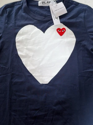 Comme des Garçons Play hjärttshirt - Mörkblå t-shirt från Comme des Garçons Play med stort vitt hjärta på bröstet och det ikoniska röda hjärtat med ögon broderat upptill. Klassisk rund hals och korta ärmar. Perfekt statement-plagg för dig som gillar streetwear och unika prints.