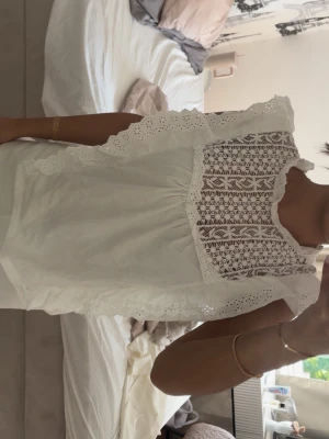 Vit spetsblus med volangdetaljer - Söt vit blus med spets upptill och volangdetaljer längs axlarna. Blusen är ärmlös och har ett romantiskt mönster i spetsen framtill. Perfekt för en trendig och feminin look. Materialet känns lätt och luftigt.
