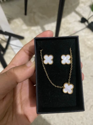 Blomformad smyckesset i guld och vitt - Snyggt smyckesset med halsband och örhängen i guldtonad metall. Både halsbandet och örhängena har vita, fyrklöverformade hängen med guldfärgad kant. Perfekt för dig som gillar stilrena och eleganta accessoarer.