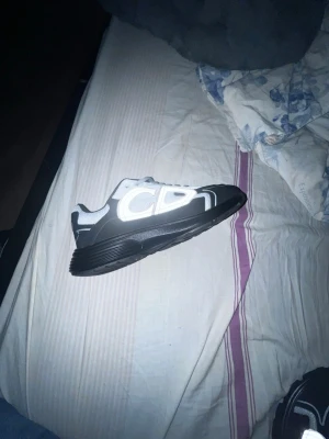 Svarta och vita CDG sneakers - Säljer ett par svarta och vita sneakers från Comme des Garçons med reflekterande detaljer och tydligt CDG-tryck på sidan. Skorna har en låg silhuett, svart sula och ovandel i mesh och syntetmaterial. Perfekta för dig som vill sticka ut med en unik design.