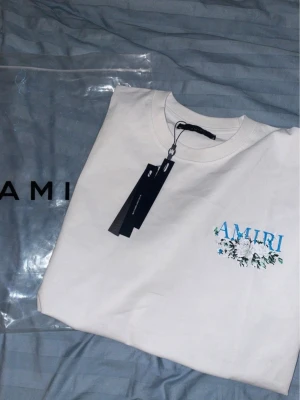 Vit AMIRI t-shirt med blommor - Säljer en fräsch vit t-shirt från AMIRI med blått och grönt blommotiv och logga på bröstet. Klassisk rund hals och korta ärmar. Tillverkad i mjuk bomull för skön känsla. Perfekt för dig som gillar stilrena plagg med en twist.               98% Bomull 2% Elastan