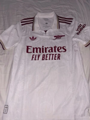 Vit Arsenal matchtröja authentic  - Säljer en vit Arsenal matchtröja från Adidas med vinröda detaljer och loggor. Tröjan har klassisk krage, korta ärmar och tryck som 'Emirates Fly Better' och 'Visit Rwanda'. Perfekt för fotbollsfans som vill sticka ut på läktaren.