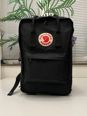 Fjällräven Kånken Laptop 15" ryggsäck, svart - Svart ryggsäck med ett stort huvudfack som stängs med dragkedja. Väskan har ett separat vadderat fack för en 15-tums laptop. Det finns en mindre ficka med dragkedja framtill och öppna sidfickor. Ryggsäcken har justerbara axelremmar och handtag upptill som kan fästas ihop med en tryckknapp. Inuti finns en uttagbar sittdyna. *** använt flera gånger nästan ny.  Höjd:  38 cm Bredd:  26 cm Djup:  17 cm Volym:  18 l Laptopfack:  Ja Storlek på laptop:  15” Mått på laptop:  36 x 26 x 3 cm Vikt:  460 g 