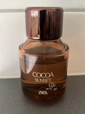 Zara Cocoa Sunset parfym - En stilren parfymflaska från Zara med doften Cocoa Sunset. Flaskan är rundad och gjord i transparent brunt glas med en blank, mörkbrun kork. Texten på flaskan är vit och designen känns modern och trendig. Perfekt för dig som gillar varma, söta dofter.