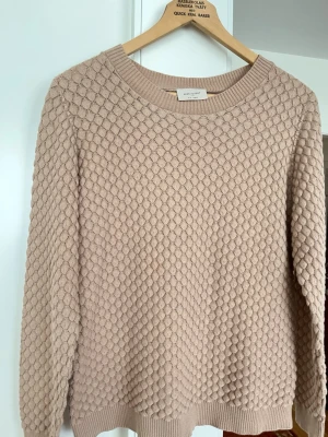 Beige stickad tröja från Freequent - Säljer en beige stickad tröja från Freequent med ett unikt bubbligt strukturmönster. Tröjan har rund halsringning, långa ärmar och ribbade muddar vid ärmslut och nederkant. Perfekt för dig som gillar en stilren men ändå intressant look.