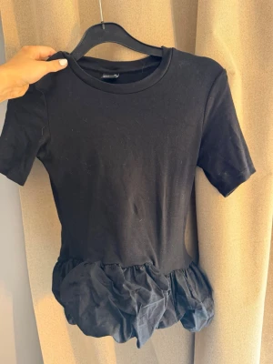 Svart topp med volangdetalj - Svart topp med korta ärmar och rund halsringning. Nedtill finns en unik volangdetalj som ger toppen en cool och trendig look. Perfekt för dig som vill sticka ut lite extra. Materialet känns mjukt och bekvämt mot huden.