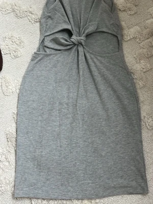 Grå ribbad klänning med knyt - Snygg grå klänning med ribbad struktur och knyt framtill som skapar en cool cut-out detalj. Klänningen är ärmlös och har en kroppsnära passform som framhäver siluetten. Perfekt för dig som gillar trendiga och unika plagg.