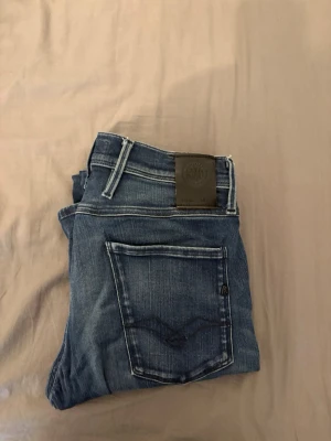 Blå jeans från Replay - Säljer ett par snygga blå jeans från Replay med klassisk femficksmodell och coola slitningar. Jeansen har kontrastsömmar, läderpatch bak i midjan och raka ben. Perfekta för dig som gillar en avslappnad men stilren look.
