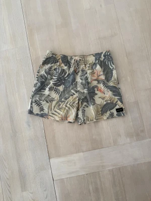 Mönstrade badbyxor - Säljer ett par snygga badbyxor med tropiskt mönster i beige, grönt och orange. Shortsen har elastisk midja med dragsko, sidofickor och en bakficka med lock. 