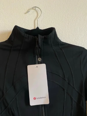 Svart träningsjacka från Lululemon - Slimmad svart träningsjacka från Lululemon med hög krage, hel dragkedja och två fickor med dragkedja. Tillverkad i stretchigt funktionsmaterial som andas och formar sig efter kroppen. Perfekt för gymmet eller löprundan.