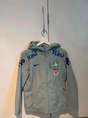 Brasilien windbreaker  - Ljusgrön vindjacka från Nike med huva och dragkedja. Jackan har Brasilien fotbollslandslagets logga på bröstet, samt tryck i blått och grönt från sponsorer som Itaú och Vivo. Två sidofickor med dragkedja och justerbar huva. Perfekt för sportiga dagar. Den är storlek L men passar perfekt för folk som har storlek M